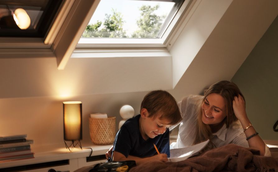 Boy reading up from a book to his mother in his bedroom where there are two VELUX roof windows behind them, which got blackout blinds.