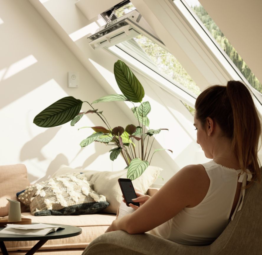 Woman controlling her VELUX roof windows in her living room with the VELUX ACTIVE app on her smartphone.