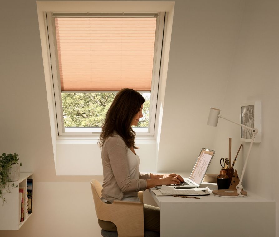 Woman sitting in her modern home office writing an email on her labtop with a VELUX roof window behnd her, which has a beige awning.