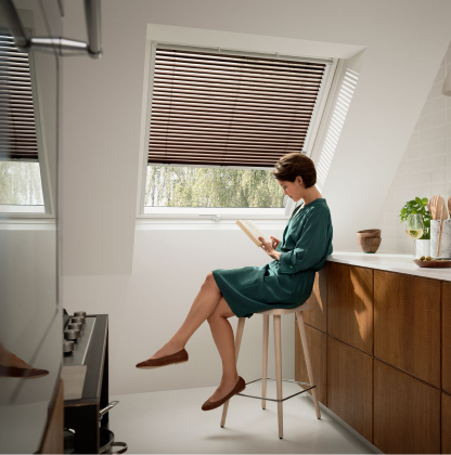 Woman sitting and reading a magazine, in her modern kitchen with one big VELUX roof window behind her, which has a purple VELUX awning blind.