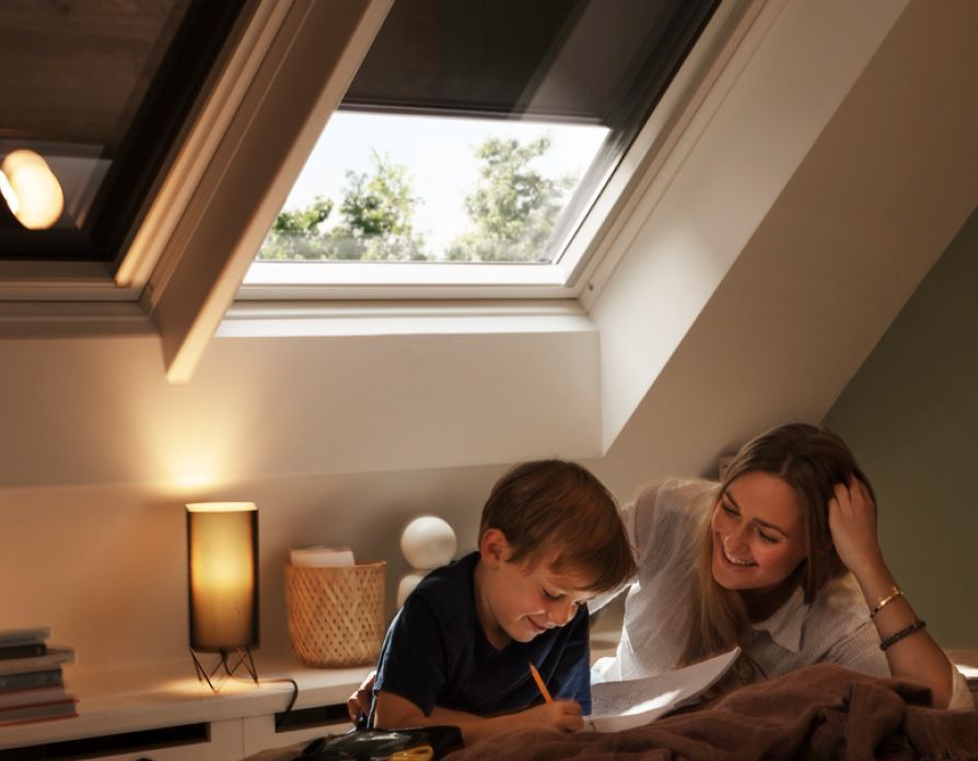 Boy reading up from a book to his mother in his bedroom where there are two VELUX roof windows behind them, which got blackout blinds.