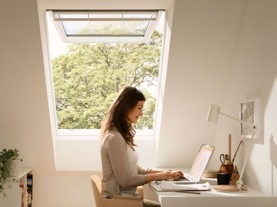 Woman sitting in her modern home office writing an email on her labtop with a VELUX roof window behind her, which is open to get fresh air.