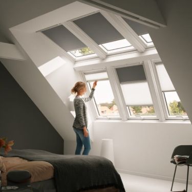 A woman looks out of her VELUX roof window, which is partially covered by Duo Blackout blinds.